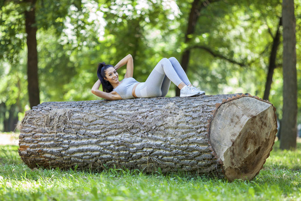 Young fit beautiful girl doing sit ups on a tree log outdoor on a grass field in forest during summer sunny day - Photo, Image