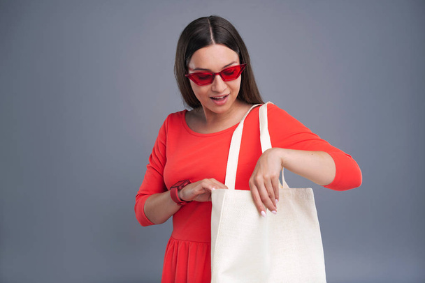 Fashion icon. Attractive dark-haired woman in a red dress wearing red glasses and checking the content of her tote bag - Fotoğraf, Görsel