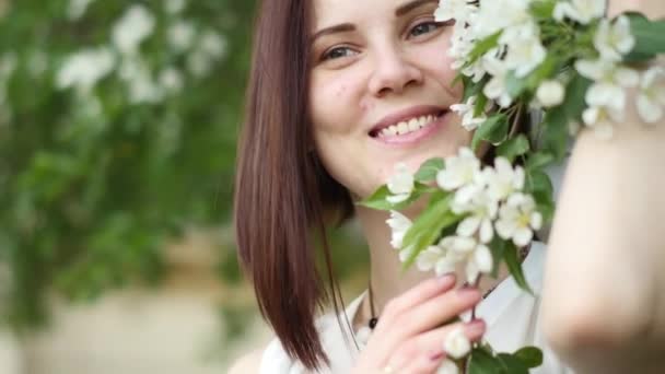 Beautiful girl portrait near a flowering tree in the park. The concept of youth and natural beauty. A young girl smells like the apple tree in the spring in the garden - Filmagem, Vídeo