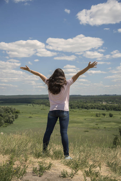 Young hipster girl is travelling in summer holidays. Woman in modern dress is standing on the hill and looking at the field. Blue sky and white clouds. Freedom and lifestyle concept. Beautiful nature - Φωτογραφία, εικόνα