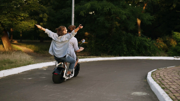 Young, happy cheerful couple riding a minibike in the parkside. Girl is feeling great, outstretched hands. Backside view - Footage, Video