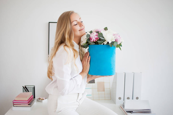 Stylish young blonde in office, white shirt and pants. Holds a luxurious bouquet of peonies in a blue hat box. - Foto, Imagen
