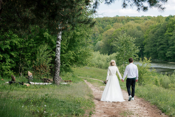 Beautiful bride and groom go holding hands in the wood near river, back view - Foto, immagini