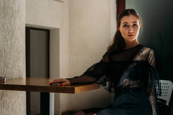 beautiful brunette sitting on a white chair at a wooden table. on it is a black dress with a platen and a star on it. waiting for a meeting. Positioning. female portrait. cafe in the city - Photo, Image