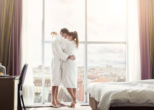 Νεαρό ζευγάρι με αγάπη στο δωμάτιο του ξενοδοχείου το πρωί, στέκεται στο παράθυρο - Φωτογραφία, εικόνα