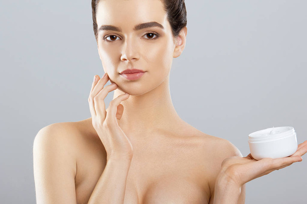 Woman Face Skin Care. Portrait Of Attractive Young Female Applying Cream  And Holding Bottle. Closeup Of Smiling Girl With Natural Makeup And Fresh Skin. Beauty Cosmetics.  - 写真・画像