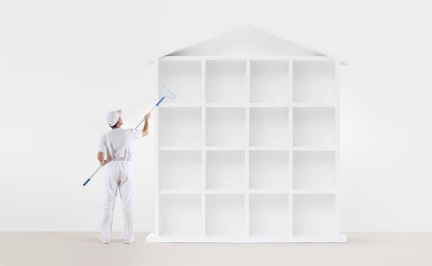 Home Service Konzept. Maler Mann mit Farbroller, der ein weißes Hausmodell isoliert auf den leeren Wandhintergrund malte - Foto, Bild