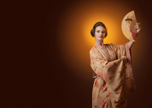 Joven hermosa mujer en kimono tradicional japonés bailando con un ventilador. Cabello y maquillaje perfectos. Espacio libre para tu contenido
. - Foto, Imagen