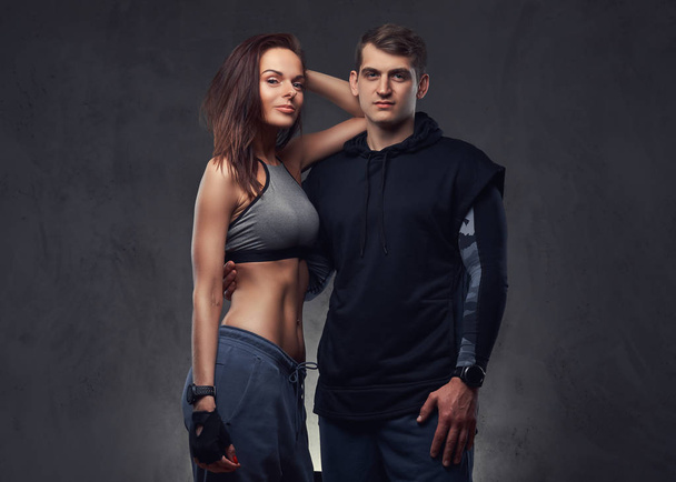 Attractive couple, slim brunette female and handsome guy in sportswear cuddling in a studio. Isolated on a dark background. - Foto, afbeelding