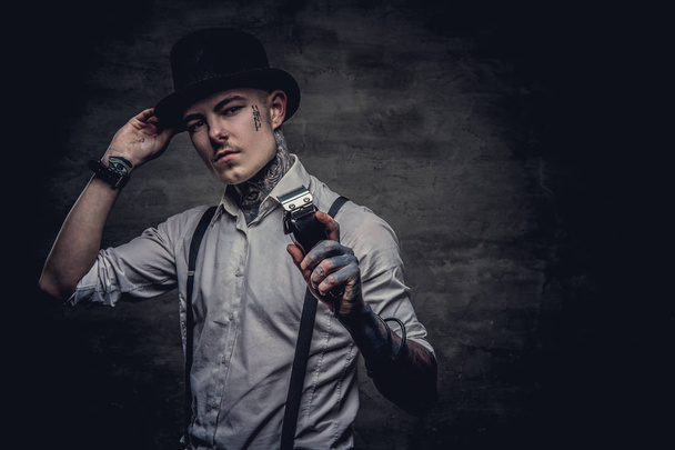 Retrato de um cabeleireiro tatuado antiquado vestindo uma camisa branca com suspensórios e chapéu de cilindro, olhando para uma câmera, segura um aparador. Isolado em um fundo texturizado escuro
. - Foto, Imagem