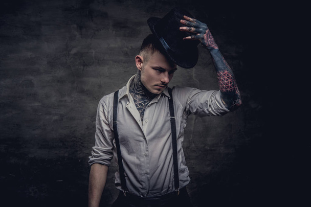 Retrato de un joven tatuado a la antigua con camisa blanca y tirantes sostiene un sombrero de cilindro. Aislado sobre fondo oscuro
. - Foto, imagen