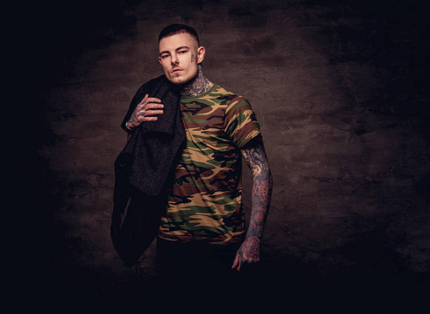 Un joven tatuado con una camiseta militar sostiene una pose de chaqueta negra en el estudio. Aislado sobre fondo de textura oscura
. - Foto, Imagen