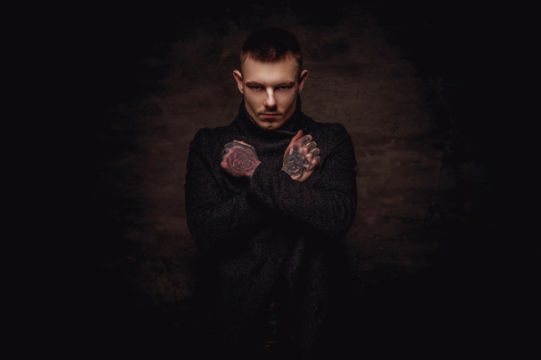 Tetovaný mladý muž v černém kabátku pózuje s překřížením rukou ve studiu. Izolované na tmavý podklad s texturou. - Fotografie, Obrázek
