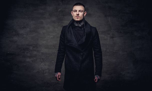 Tetovált fiatal srác egy fekete kabát, pózol a stúdióban. Egy sötét textúrázott háttérre elszigetelt. - Fotó, kép