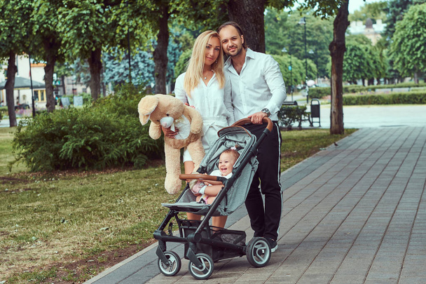 Feliz concepto de familia joven. Hermosa madre rubia y padre con barba elegante caminando en el parque con su hija pequeña en un cochecito de bebé
. - Foto, imagen