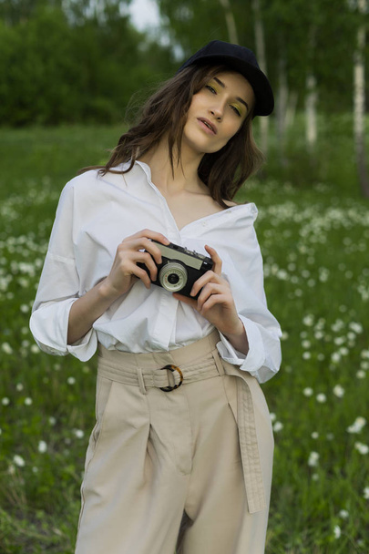 Beautiful girl photographer wearing a white shirt, beige trousers poses outdoors with a vintage camera against a background of meadow full of wild flowers. Lifestyle and commercial design. Copy space. - Valokuva, kuva