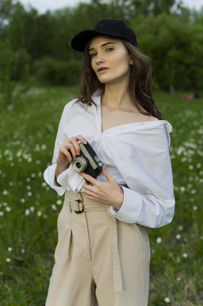 Beautiful girl photographer wearing a white shirt, beige trousers poses outdoors with a vintage camera against a background of meadow full of wild flowers. Lifestyle and commercial design. Copy space. - Photo, image