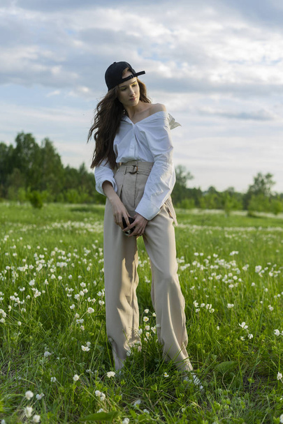 Beautiful girl photographer wearing a white shirt, beige trousers poses outdoors with a vintage camera against a background of meadow full of wild flowers. Lifestyle and commercial design. Copy space. - Foto, imagen