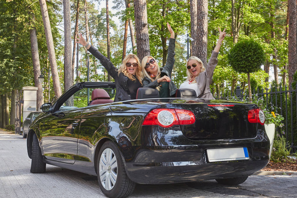 Joyful fashionable female friends raise their hands while sitting in luxury cabriolet car in a park. - Photo, Image