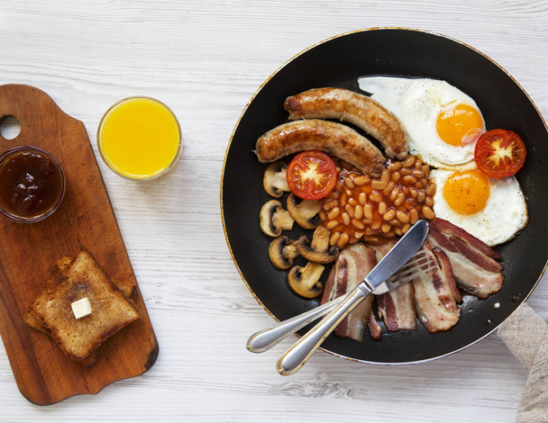 Café da manhã inglês completo em uma panela com ovos fritos, bacon, feijão, salsichas e torradas em um fundo de madeira branca, vista superior. Por cima, por cima.
. - Foto, Imagem