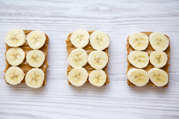 Tostadas veganas con mantequilla de maní y plátano sobre un fondo de madera blanca, desde arriba
. - Foto, Imagen