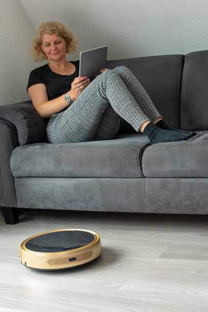 Woman is steering her vacuum cleaning robot from the sofa - Photo, Image