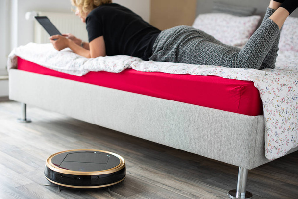 Vacuum cleaning robot with relaxed woman reading in the background - Photo, Image