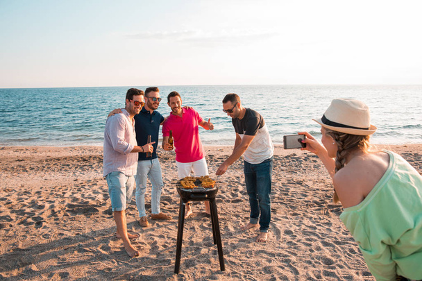Woman is making a photo to  a group of men that are preparing a barbecue on the beach. Vacation concept. - Photo, Image