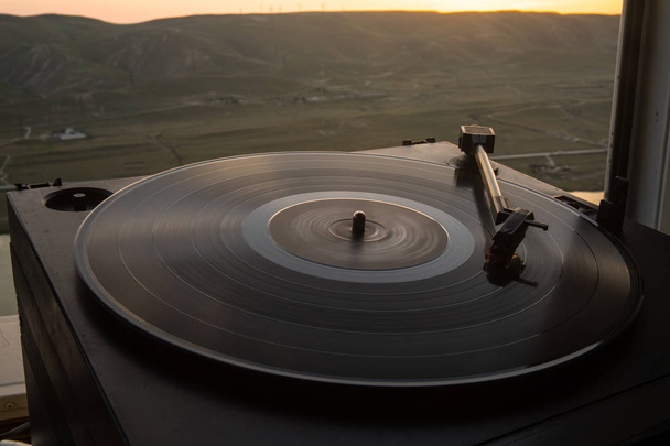 Πικάπ βινυλίου πικάπ στο φόντο ένα ηλιοβασίλεμα πάνω από τα βουνά. Τεχνολογία ήχου για Dj για να αναμίξετε & παίζουν μουσική. Μαύρο δίσκο βινυλίου. Πικάπ vintage βινύλιο. Βελόνα σε έναν δίσκο βινυλίου - Φωτογραφία, εικόνα
