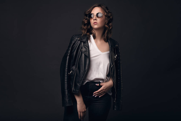 A young brunette girl with a stylish hairstyle on a dark background in a leather jacket, white T-shirt and sunglasses. Professional model posing on an isolated dark background with space for text - Fotografie, Obrázek