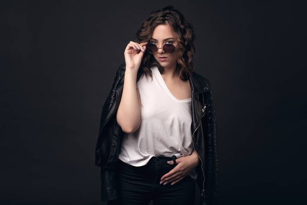 A young brunette girl with a stylish hairstyle on a dark background in a leather jacket, white T-shirt and sunglasses. Professional model posing on an isolated dark background with space for text - Valokuva, kuva