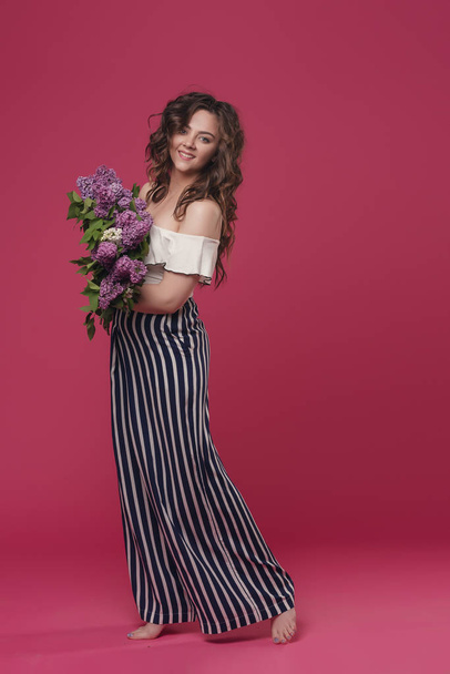 Young beautiful girl with long wavy hair with a bouquet of purple flowers happy hurrying on a date. On a pink isolated background with space for text - Zdjęcie, obraz