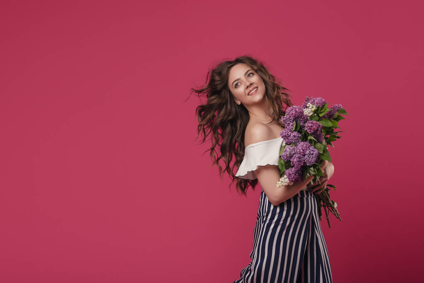 Young beautiful girl with long wavy hair with a bouquet of purple flowers happy hurrying on a date. On a pink isolated background with space for text - Photo, Image