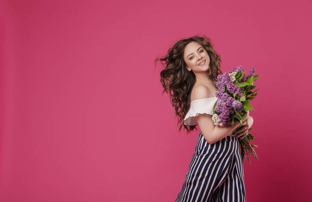 Young beautiful girl with long wavy hair with a bouquet of purple flowers happy hurrying on a date. On a pink isolated background with space for text - Foto, afbeelding