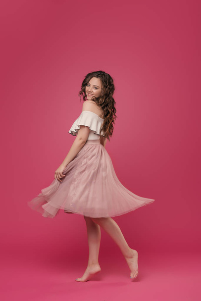 Young stylish girl in pink light skirt and white T-shirt on pink isolated background with place for text. The girl cheerfully dances and jumps like a ballerina - Fotó, kép