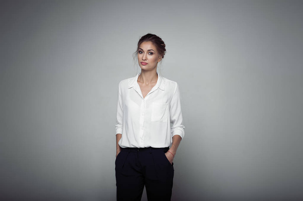 Interested businesswoman with trendy makeup posing on gray background in studio. Indoor photo of serious young lady in white blouses classic black pants standing in confident pose. - Valokuva, kuva