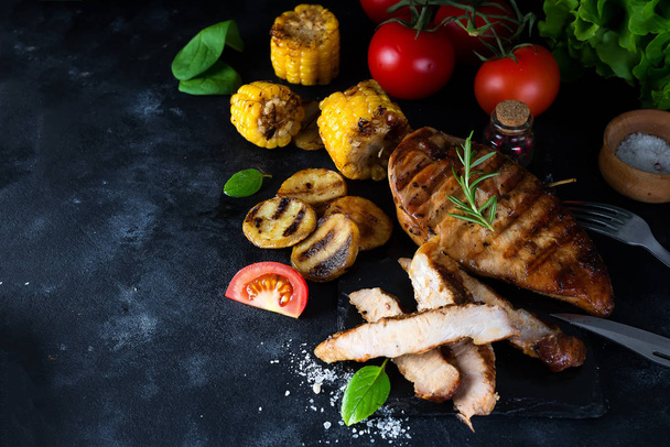 Pine slice and chicken grill steaks, tomatoes, corn and potatoes on a grill with basil and rosemary on a black concrete background. Flat lay with copy space - Photo, Image