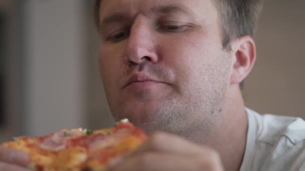 de man is het eten van pizza. Op een dikke vent, een wit T-shirt. Hij houdt van een stukje pizza in zijn handen en eet met een eetlust - Video