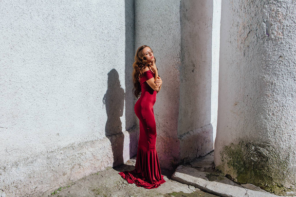 Beautiful young woman in a long red evening gown with a train standing next to the old wall - Valokuva, kuva