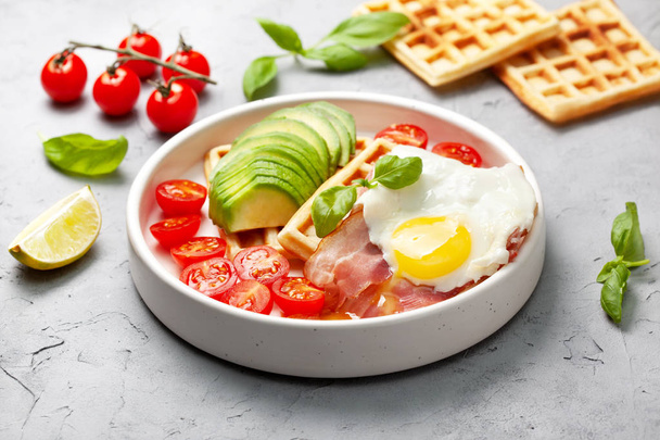 healthy breakfast. Belgian waffles with avocado, bacon and fried egg in a white plate on a gray concrete background. - Photo, image