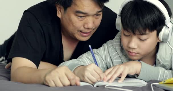 アジアの父の前面からドリー撮影彼の若い息子を助けるは、少年は自宅に彼の宿題をしている間 - 映像、動画