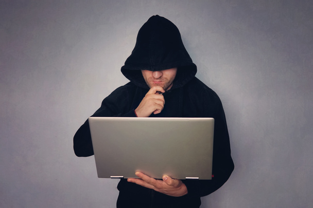 cybercriminaliteit, doordachte hacken en technologie misdaad - mannelijke hacker in donkere kamer met laptopcomputer, hacker in een zwarte kap met een grijze moderne ultrabook, diefstal van identiteit. - Foto, afbeelding