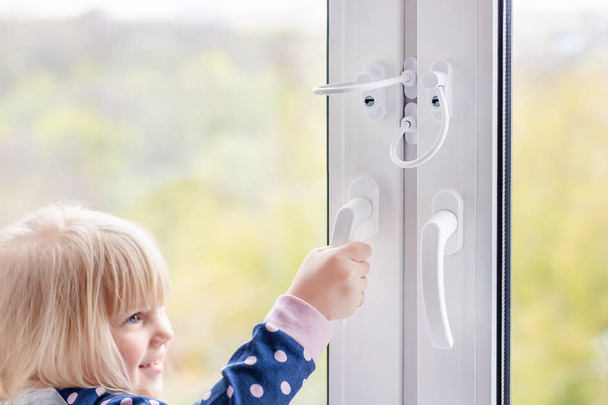 Little cute toddler girl trying to open window in apartment at high-tower building. Children window protection lock. Cable safety guard prevent opening window by child. Prevention of falling accident - Zdjęcie, obraz
