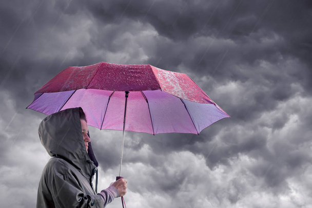 Woman with an umbrella against the background of a dark stormy sky. The woman is hiding under an umbrella from the rain. Protection from danger, natural disasters - Photo, Image