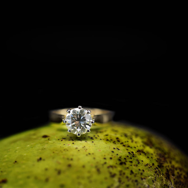 A large diamond solitaire set into a yellow gold ring, resting on a green mango against a black background - Photo, Image