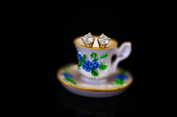 Um par de brincos de diamante cravejado de ouro amarelo, sentado em uma xícara de chá floral em miniatura contra um fundo preto
 - Foto, Imagem