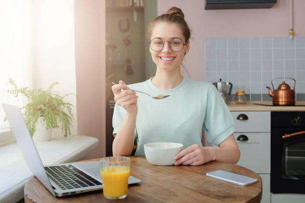 Indoors shot of beautiful young smiling woman having delicious tasty breakfast, sitting at wooden table in modern colorful kitchen - Zdjęcie, obraz