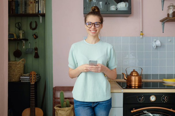 Young smiley woman standing in the colorful kitchen, holding her phone with both hands, looking straight to the camera - Foto, immagini