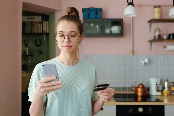 Closeup shot of young smiley woman wearing trendy round eyeglasses, paying with credit card while e-shopping, using mobile app, buying new dress. Online transaction with digital currecny concept - Foto, afbeelding