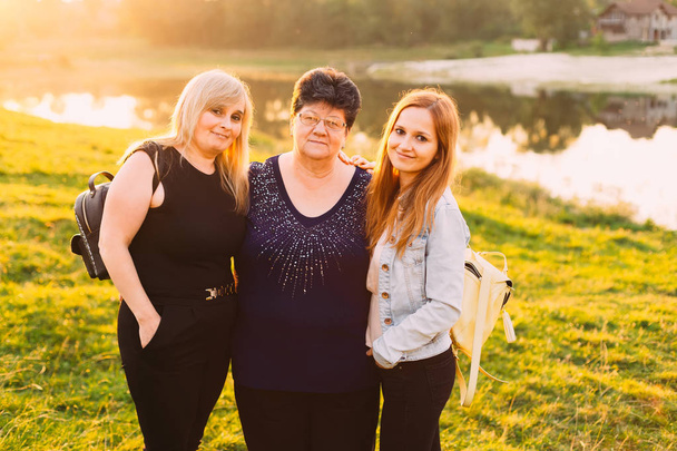 Babcia, matka i córka przyjęli i patrząc w obiektyw aparatu na tle zachodu słońca i jezioro. trzy pokolenia w jedno zdjęcie - Zdjęcie, obraz